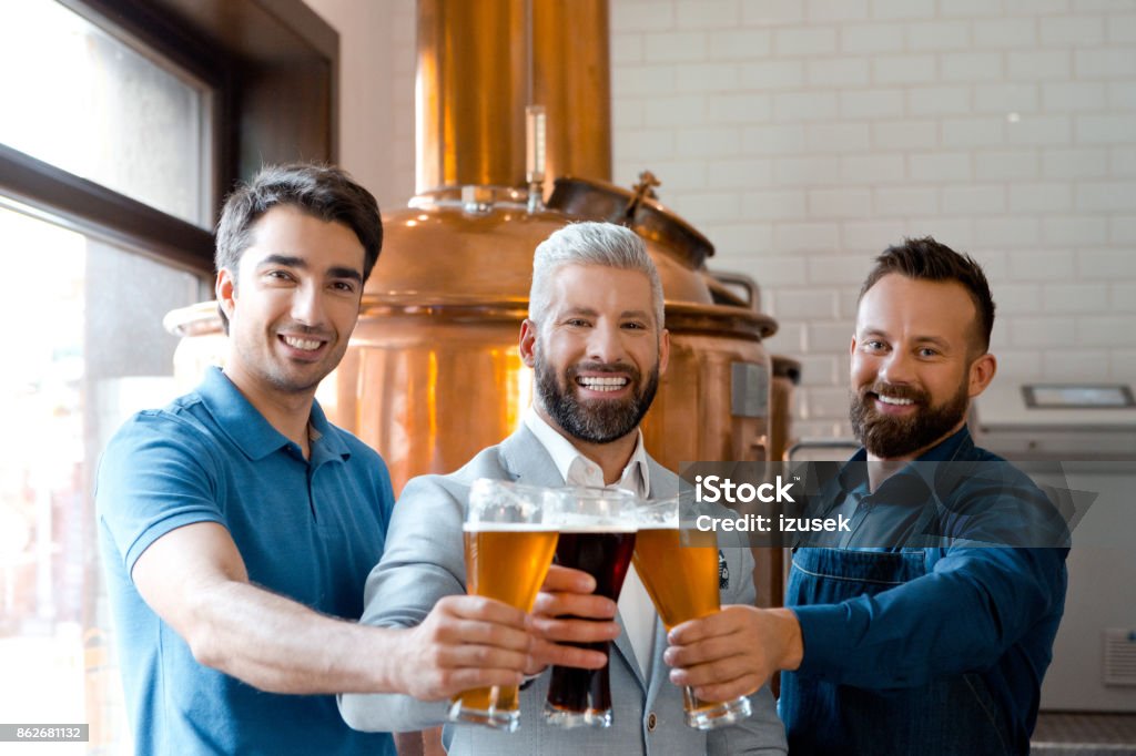 Microbrewery team toasting with beer Owner, brewer master and bartender toasting with beer glasses in microbrewery, smiling at the camera. Three People Stock Photo