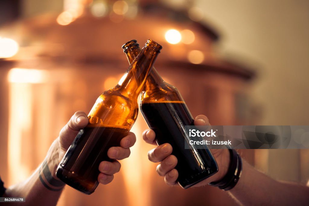 Friends toasting with beer bottles in the microbrewery Friends toasting with beer bottles in the microbrewery. Close up of bottles and hands, unrecognizable people. Copper vat in the background. Celebratory Toast Stock Photo