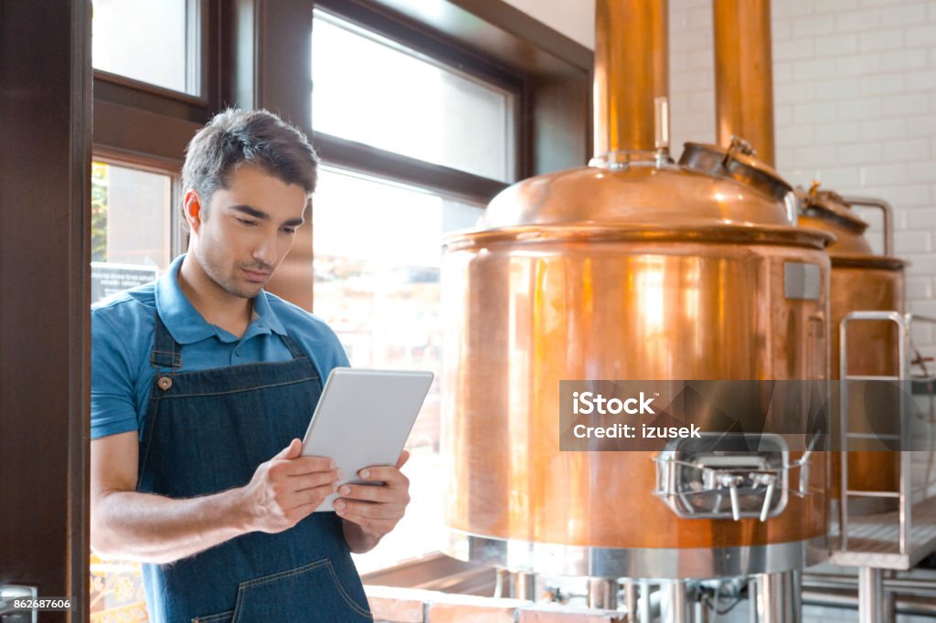 Bartender using a digital tablet in micro brewery Bartender standing in the pub and using a digital tablet. Copper vats in the background. Brewery Stock Photo