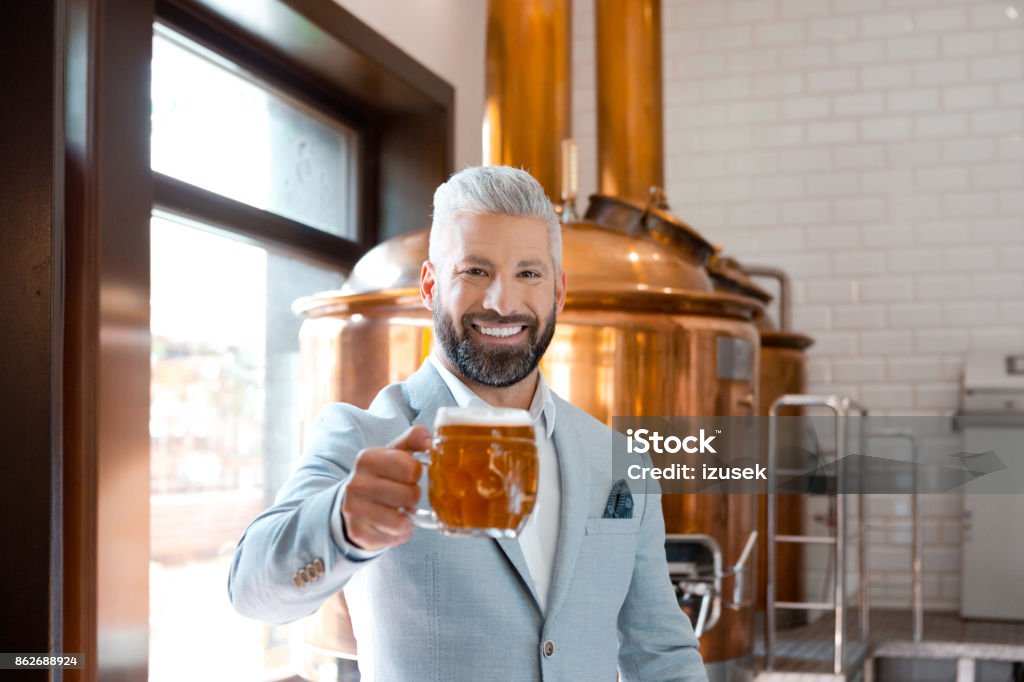 Happy elegant man holding a beer mug in microbrewery The microbrewery owner standing in front to copper vat and holding beer mug, toasting at the camera. Selective focus. Adult Stock Photo