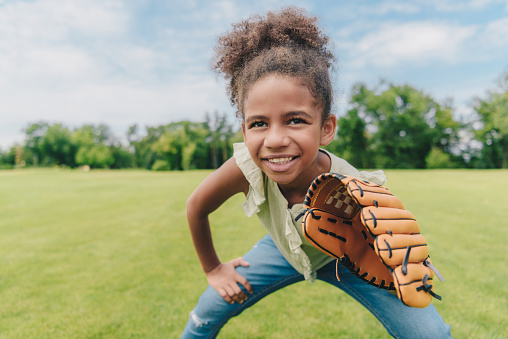 portrait of smiling african american little girl with baseball glove playing baseball in park