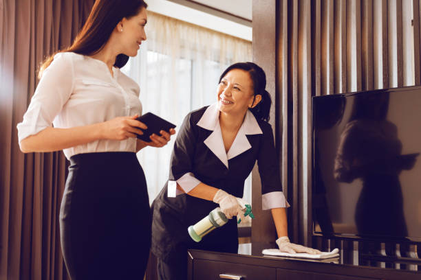Positive delighted chambermaid polishing furniture Friendly conversation. Attractive hotel guest standing in semi position and holding tablet in both hands while looking at room cleaner housekeeping staff photos stock pictures, royalty-free photos & images
