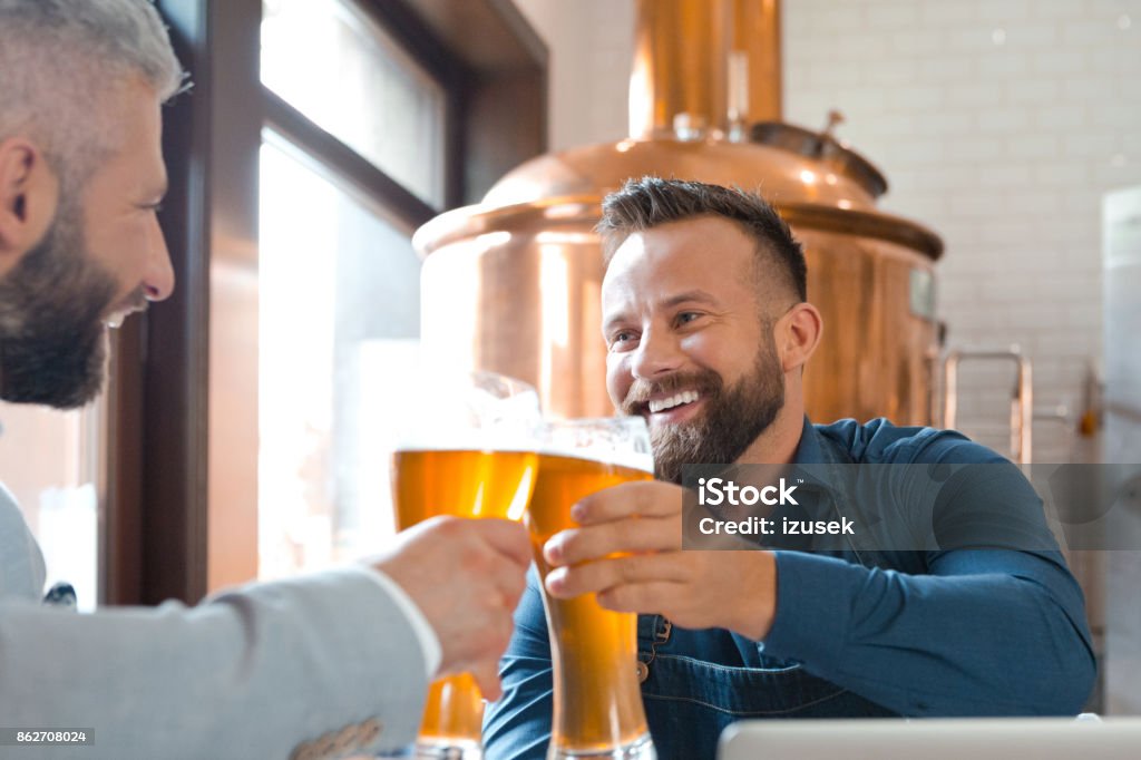 Brewer master toasting with beer glass with his friend Brewer master toasting with beer glass with his friend. Copper vat in the background. Brewery Stock Photo