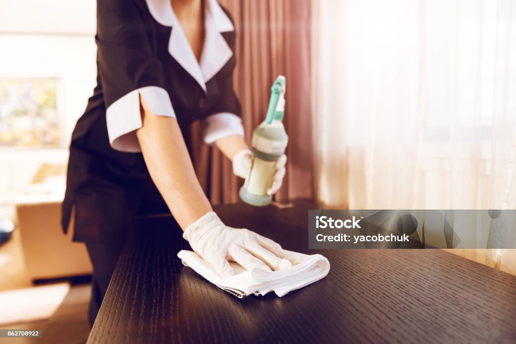 Close up of female hand that using serviette With care. Professional maid holding cleaning liquid in left hand and standing close to chest of drawers while polishing it Hotel Stock Photo
