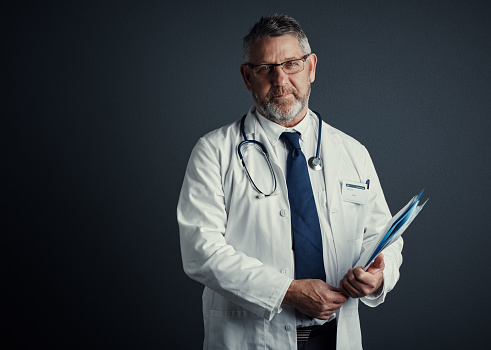 Studio portrait of a handsome mature male doctor holding medical records while standing against a dark background