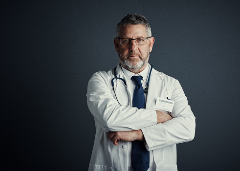 Studio portrait of a handsome mature male doctor standing with his arms folded against a dark background