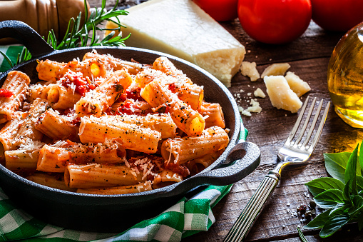 Rigatoni pasta served in a small cast iron pan with tomato sauce on rustic wooden table. Some ingredients for preparing and serving pasta like pepper shaker, olive oil, fresh tomatoes, Parmesan cheese, basil leaves, rosemary,  salt, pepper, and garlic are beside the plate. DSRL studio photo taken with Canon EOS 5D Mk II and Canon EF 100mm f/2.8L Macro IS USM