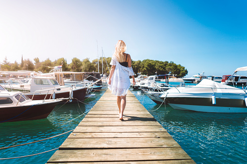 Young woman in the summer, walking on a wooden dock, pier in the marina, boats and ships in the harbour, nautical vessel, fit and beautiful young woman in a white dress
