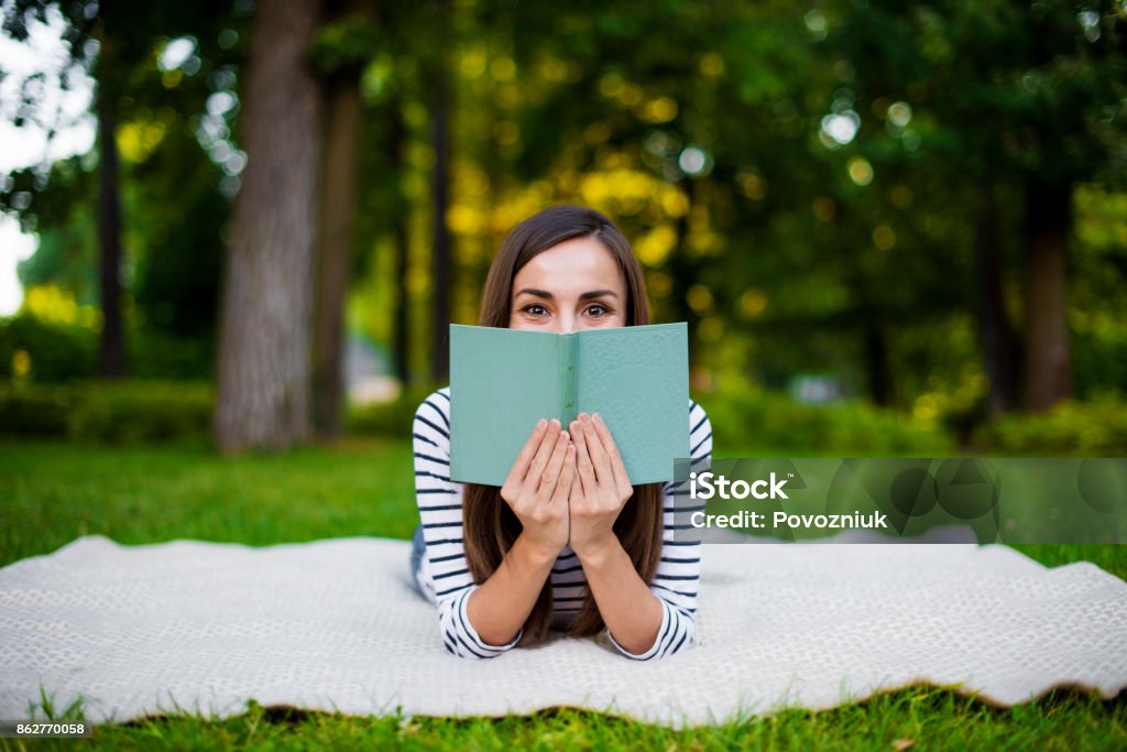 Beautiful smiling modern young girl student in casual clothes reading a book lying on a plaid in a city park Reading Stock Photo