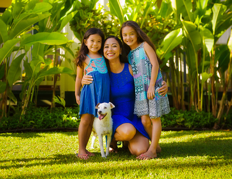 Portrait of a Hawaiian Polynesian family outdoor in their backyard. Two young girls with their mother and family pet dog, smiling and looking at camera full body group portrait.