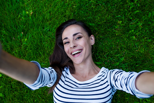 Cheerful smiling young modern woman with chic hair makes selfie on her phone while lying on green grass. View from above