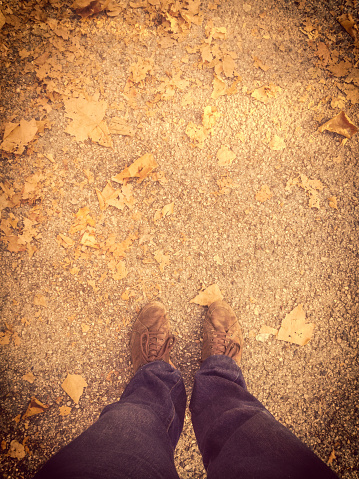 Close-up of men legs with jean pants, on gravel texture during sunny autumn season with dried leaves on ground. Vertical composition mobile camera stock photography with large copy space on top.