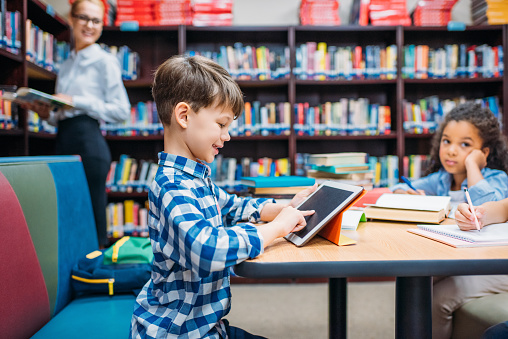 cute smiling schoolboy using tablet at library