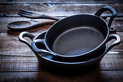 Two black cast iron pans arranged in a pile shot on rustic wooden kitchen table. An old wooden spoon and fork are behind the pans. Predominant colors are brown and black. Low key DSRL studio photo taken with Canon EOS 5D Mk II and Canon EF 100mm f/2.8L Macro IS USM