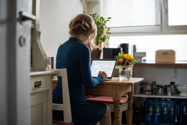 blonde jeune femme assise à la table dans une cuisine domestique, tout en travaillant sur un ordinateur portable - rear view women back back of head photos et images de collection