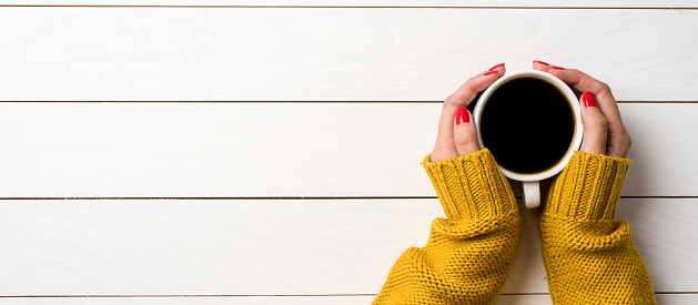 Female hands in warm sweater holding cup of coffee. Top view