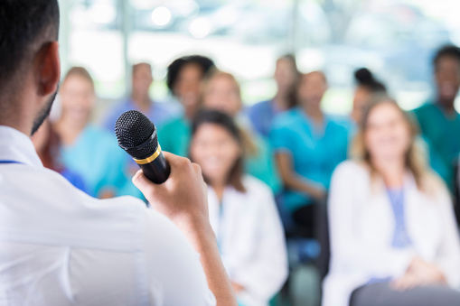 Unrecognizable healthcare professional addresses colleagues during medical conference. Focus is on the healthcare professional holding a microphone. The audience is blurred in the foreground.