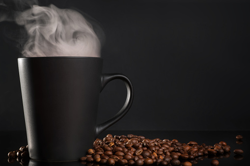 Black cup of coffee with steam and coffee beans on black table