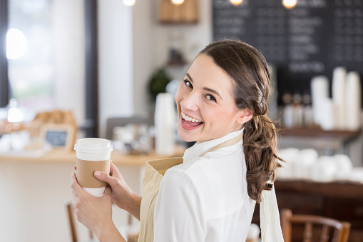 Cheerful young female waitress smiles while holding a disposable coffee cup. She is looking over her shoulder.