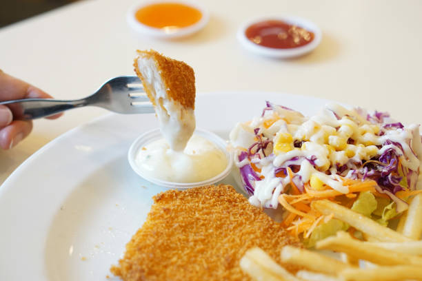 Hand holding crispy fried fish dipping in tartar sauce served with french fries.
