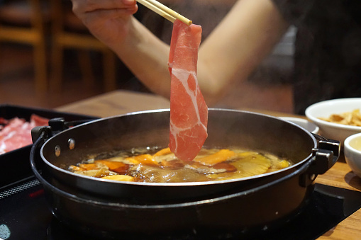 Hand holding raw pork using chopsticks on hot pot with smoke for Japanese hot pot also known as Shabu Shabu or Sukiyaki is Japanese style.