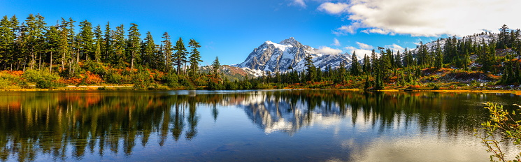 Aerial View of the bright Blue Ann Lake with an Island between Rising Mountains, covered with a lush bright green Pine Tree Forest, under a dramatic, moody, cloudy sky. Ann Lake is near the Pacific Crest Trail, North Cascades National Park in Washington, USA