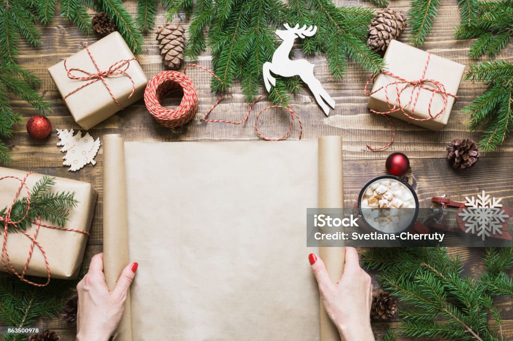 Christmas composition. Female holding empty scroll wishlist for Santa Claus or holiday cheers laid on a wooden table with christmas giftbox and decor. Flat lay, top view, copy space. Christmas Stock Photo