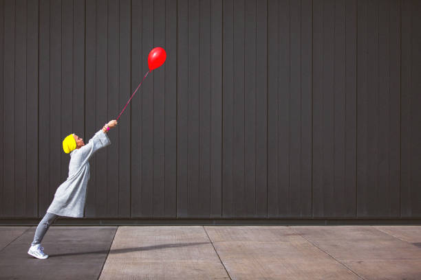 Young woman dancing and holding red balloon against the grey wall stock photo