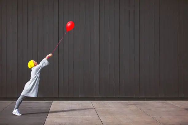 Young woman dancing and holding red balloon against the grey wall