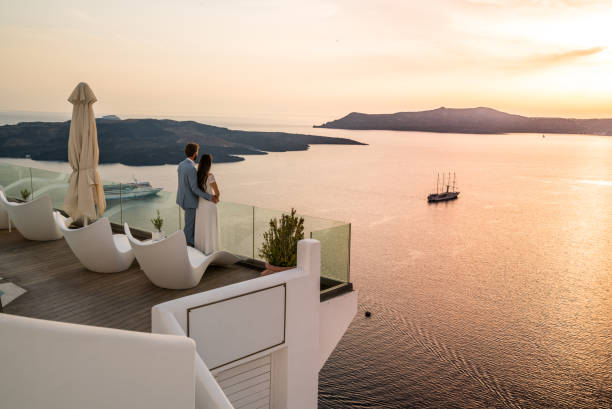 Authentic Wealth - rich couple standing on terrace with amazing sea view couple in love on romantic location - luxury hotel in greece overlooking the caldera of greek island santorini terrace with seaview vacation honeymoon high society photos stock pictures, royalty-free photos & images