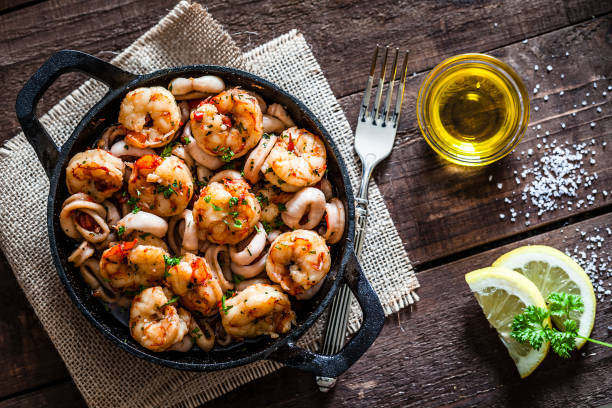 Shrimps and calamari rings cooked on iron cast pan Top view of an iron cast pan filled with fresh shrimps and calamari rings shot on rustic wooden table. Lime halves, olive oil, parsley leaves and garlic cloves are beside the pan places directly on the table. A fork is also on the table. Predominant colors are red and brown. DSRL studio photo taken with Canon EOS 5D Mk II and Canon EF 100mm f/2.8L Macro IS USM prawn grilled seafood prepared shrimp stock pictures, royalty-free photos & images