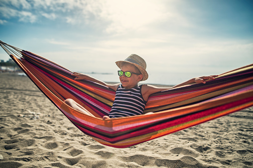 Little boy resting on beach hammock. The boy is wearing sunglasses and is taking selfie.