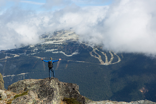 A male hiker stands on top of the mountain with his arms outstretched looking at the view