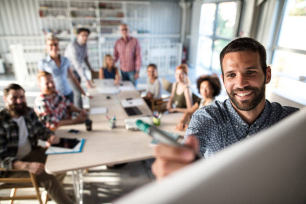 Happy businessman writing on whiteboard during business presentation in the office. Happy businessman writing a business plan on whiteboard to his team on a meeting in board room. whiteboard stock pictures, royalty-free photos & images