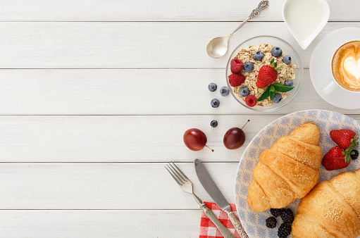 Rich continental breakfast background. French crusty croissants, muesli, lots of sweet berries and hot coffee for tasty morning meals. Delicious start of the day. Top view, copy space on white wood