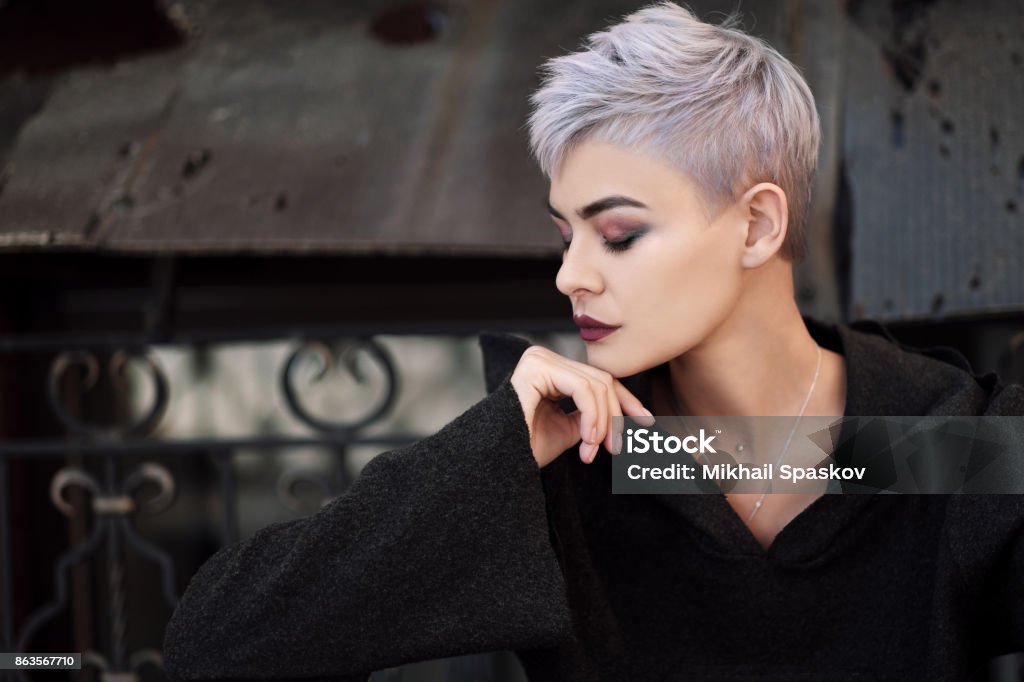 Young beautiful fashion girl shooting outdoors near brick wall at house Young beautiful girl with short gray hair in a black jacket and aggressive leather boots, fashionable shooting on the street near the brick wall of the house Short Hair Stock Photo