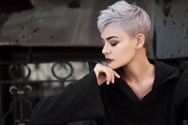 Young beautiful girl with short gray hair in a black jacket and aggressive leather boots, fashionable shooting on the street near the brick wall of the house