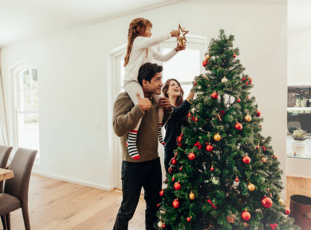 Family celebrating Christmas at home. Family decorating a Christmas tree. Young man with his daughter on his shoulders helping her decorate the Christmas tree. traditional christmas stock pictures, royalty-free photos & images