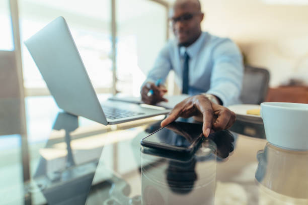 Man working on laptop computer sitting at his desk in office. Man reaching out to pick his mobile phone placed on table.