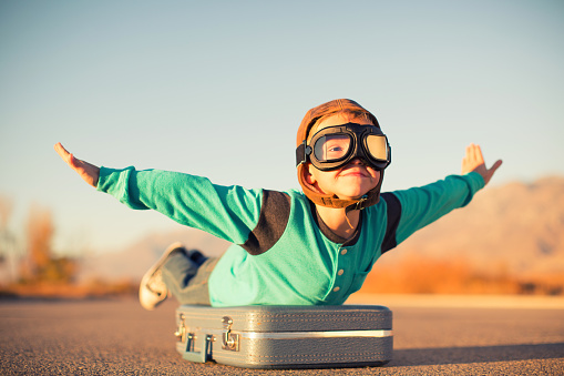 A young boy dressed in retro clothing and flying goggles dreams of flying on an exotic vacation at a far off destination. He is outstretching his arms like an airplane while on top of a suitcase and he has a happy expression on his face. Image taken in Utah, USA.