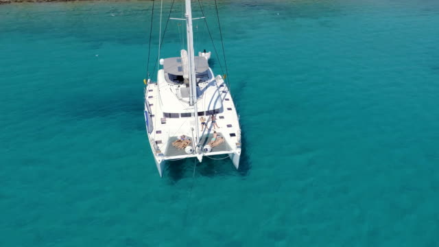 Aerial View of a Anchored Catamaran Yacht with People Sunbathing on it's Deck. Boat Stands in Azure Sea Waters With Coral Reef Visible.