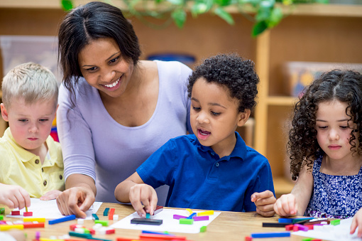 A multi-ethnic group of young children are indoors in a school. They are wearing casual clothing. They are sitting at a table and playing with blocks along with their teacher.