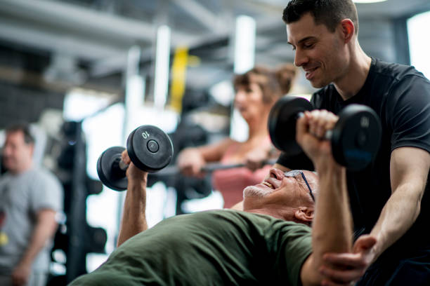 Personal Trainer A senior man and a male trainer are indoors in a fitness center. They are wearing casual athletic clothing. The man is lifting weights with the guidance of the trainer. trainer stock pictures, royalty-free photos & images