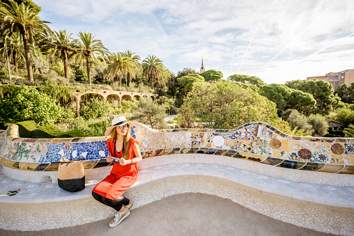 Young woman tourist in red dress sitting on the bench decorated with mosaic in the famous Guell park in Barcelona. Wide angle image with copy space