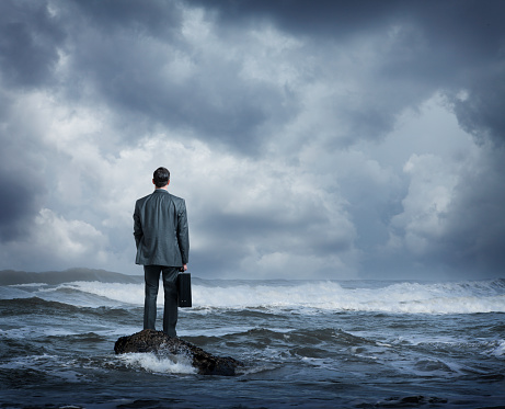A rear view of a businessman as he stands on a small rock in a rough ocean surf.  He stands holding his briefcase as he looks out across the rough waters and out towards the stormy sea.
