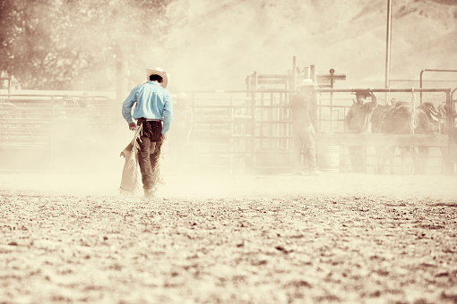 A solitary Cowboy walking back to the chutes after his ride has been completed.  Other cowboys  are muted in the dust and in the background.  Copy Space.