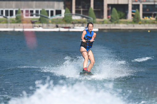 Portrait of a female wakeboarder smiling with pride because she successfully got up on the board. It is summer and there is bright sunlight. She is wake boarding on a lake and is wearing a lifejacket.