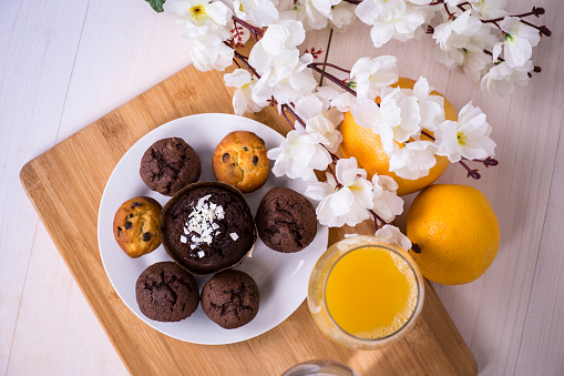 A high angle shot of muffins on a plate placed on a wooden board with flowers, two oranges and a glass of orange juice by its side.