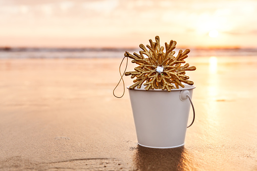 Golden Snowflake Collected in a White Bucket at Sunset on the background of Beach and Sea in California, Winter Holiday Concept