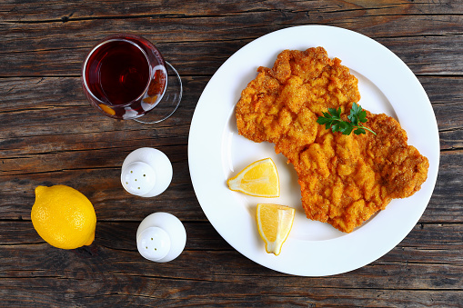 delicious golden brown classic Wiener schnitzel - or breaded veal cutlets served on white plate with lemon slices on dark wooden table with glass of red wine, horizontal view from above